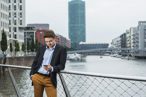 Deutschland, Hessen, Frankfurt, junger Geschäftsmann steht auf einer Brücke und benutzt sein Smartphone - UUF001830