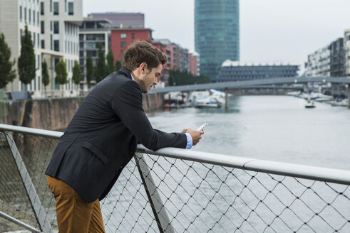 Deutschland, Hessen, Frankfurt, junger Geschäftsmann steht auf einer Brücke und benutzt sein Smartphone - UUF001832