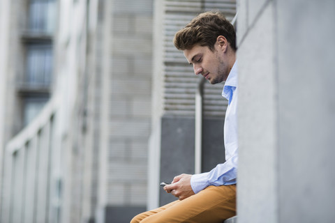 Deutschland, Hessen, Frankfurt, Porträt eines sitzenden jungen Mannes, der sein Smartphone benutzt, lizenzfreies Stockfoto