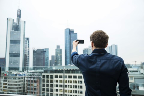 Deutschland, Hessen, Frankfurt, junger Mann fotografiert die Skyline mit seinem Smartphone, lizenzfreies Stockfoto