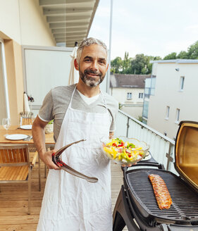 Porträt eines lächelnden Mannes mit Salatschüssel und Zange auf seinem Balkon - MBEF001283