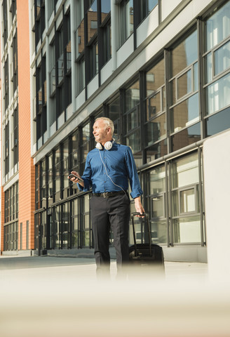 Geschäftsmann mit Koffer, Mobiltelefon und Kopfhörern vor einem Bürogebäude, lizenzfreies Stockfoto