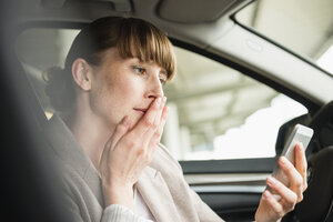 Porträt einer Geschäftsfrau, die in einem Auto sitzt und mit der Hand auf dem Mund auf ihr Smartphone schaut - FMKYF000531