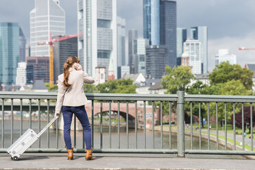 Germany, Hesse, Frankfurt, telephoning businesswoman standing with rolling suitcase on a bridge - FMKYF000560