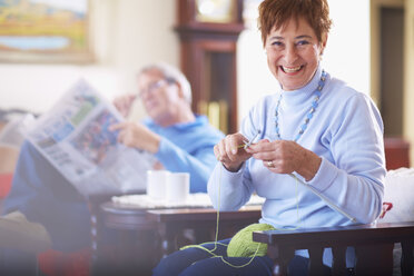 Senior woman knitting with husband in background reading newspaper - ZEF001064
