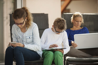 Mother and her two children relaxing with digital tablet, smartphone and laptop on the terrace - PAF001007