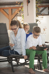 Mutter und ihre beiden Kinder entspannen sich mit Tablet, Smartphone und Laptop auf der Terrasse - PAF001011