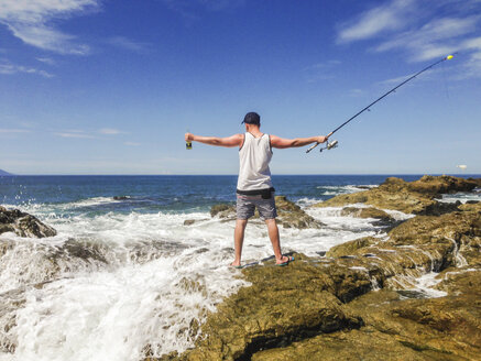Young man with fishing pole standing at the pacific coast in Mexico, waves breaking on rocky shore. - ABAF001512