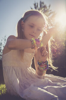 Porträt eines kleinen Mädchens, das im Sonnenlicht sitzt und viele Webstuhlarmbänder trägt - SARF000920