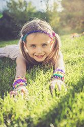 Porträt eines lächelnden kleinen Mädchens mit Webstuhlarmbändern und Haarband auf einer Wiese liegend - SARF000915