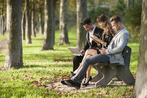 Drei Geschäftsleute sitzen auf einer Parkbank und benutzen Laptop, Smartphone und digitales Tablet, lizenzfreies Stockfoto