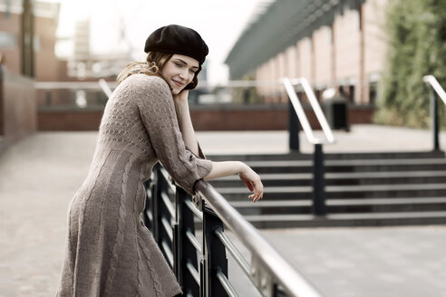 Portrait of young woman wearing beret and knitted dress leaning on a railing - GDF000501