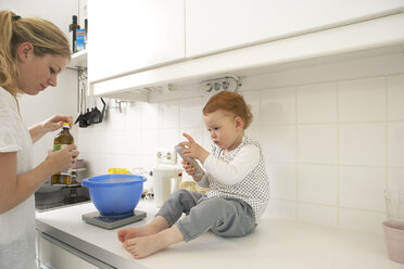 Little girl sitting on kitchen counter with smartphone while her mother baking - FSF000238