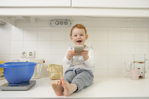 Lachendes kleines Mädchen, das mit seinem Smartphone auf dem Küchentisch sitzt, lizenzfreies Stockfoto