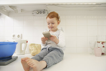 Kleines Mädchen sitzt mit Smartphone auf dem Küchentisch - FSF000248