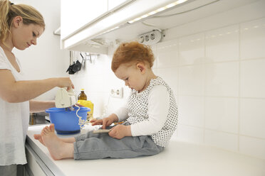 Little girl sitting on kitchen counter with smartphone while her mother baking - FSF000240