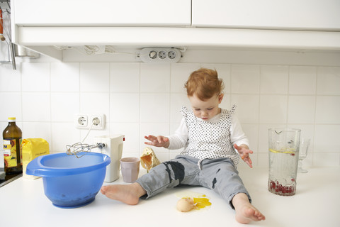 Kleines Mädchen sitzt auf dem Küchentisch und betrachtet ein zerbrochenes Ei, lizenzfreies Stockfoto