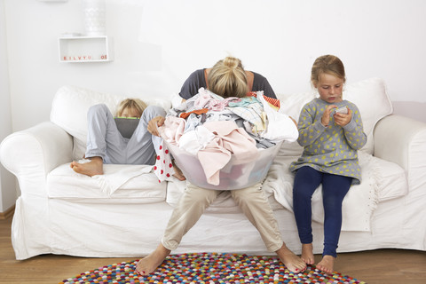 Erschöpfte Mutter mit Wäschekorb auf der Couch mit Kindern, die ein digitales Tablet und ein Mobiltelefon benutzen, lizenzfreies Stockfoto