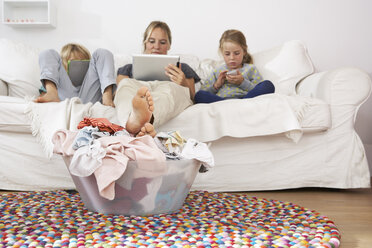 Mother, daughter and son on couch using digital tablet and cell phone with laundry basket on floor - FSF000262