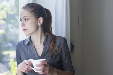 Young woman with cup of coffee looking through window - SGF000870