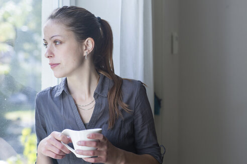 Junge Frau mit einer Tasse Kaffee schaut durch ein Fenster - SGF000870