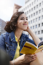 Germany, Berlin, portrait of happy young woman on city trip sitting at Potsdam Square with guidebook - FKF000728