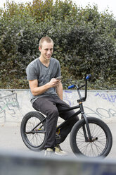 Young man with smartphone sitting on BMX bike - DRF001141