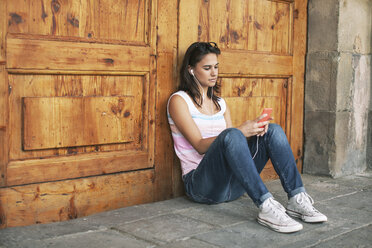 Young woman hearing music with ear phones leaning at wooden door - EBSF000326