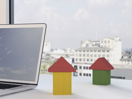 Bauklötze in Form von Häusern und Laptop auf dem Schreibtisch vor einem Fenster - UWF000211
