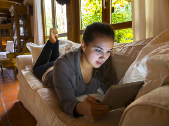 Weiblicher Teenager liegt auf der Couch im Wohnzimmer und benutzt ein digitales Tablet - AM003087