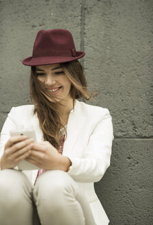 Porträt einer lächelnden jungen Frau, die vor einer grauen Wand auf ihr Smartphone schaut - UUF002353