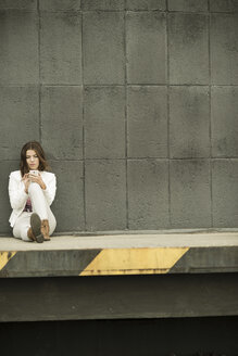 Junge Frau sitzt auf einer Rampe vor einer grauen Wand und benutzt ihr Smartphone - UUF002354