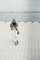 Junge Frau sitzt auf einem Poller an der Promenade und orientiert sich mit einem digitalen Tablet - UUF002356