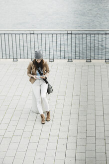 Junge Frau sitzt auf einem Poller an der Promenade und orientiert sich mit einem digitalen Tablet - UUF002356