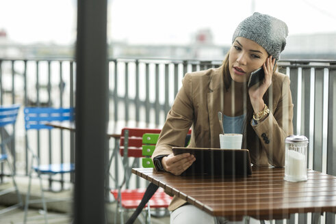 Junge Frau sitzt in einem Straßencafé und hält ein digitales Tablet, während sie mit einem Smartphone telefoniert - UUF002366