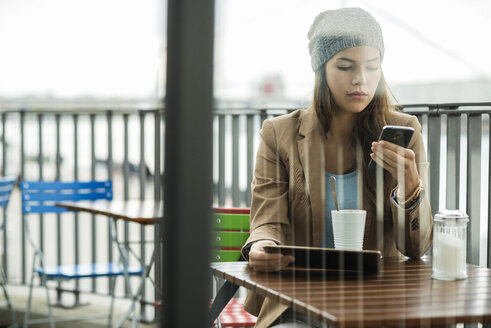 Junge Frau sitzt in einem Straßencafé und hält ein digitales Tablet, während sie ein Smartphone benutzt - UUF002367