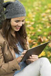 Lächelnde junge Frau lehnt an einem Baum und benutzt ein digitales Tablet - UUF002382