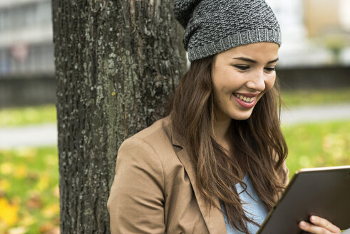 Porträt einer lächelnden jungen Frau, die sich mit einem digitalen Tablet gegen einen Baum lehnt - UUF002383