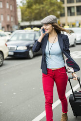 Young smiling woman with smartphone and wheeled luggage walking along a street - UUF002384