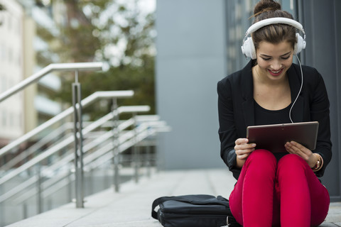 Junge Frau mit Kopfhörern, die auf einer Treppe sitzt und ein digitales Tablet benutzt, lizenzfreies Stockfoto