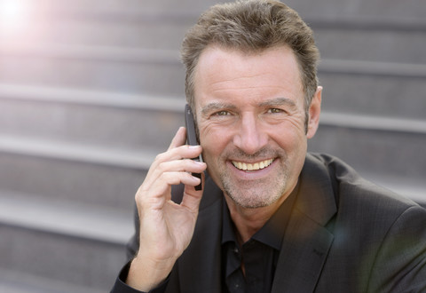 Portrait of smiling businessman sitting on steps telephoning with smartphone stock photo