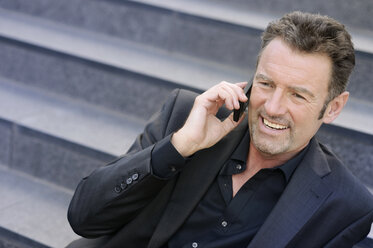 Porträt eines lächelnden Geschäftsmannes, der auf einer Treppe sitzt und mit einem Smartphone telefoniert - GUFF000041