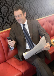 Businessman sitting in a cafe with newspaper using smartphone - GUFF000045