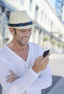 Portrait of smiling mature man with stubble and summer hat using smartphone - GUFF000051