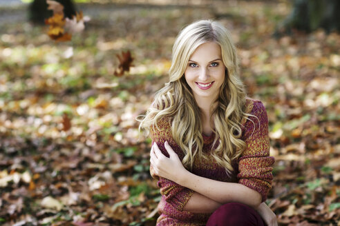 Portrait of smiling blond woman wearing knit pullover sitting in autumnal forest - GDF000540