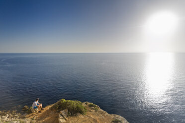 Spain, Balearic Islands, Majorca, one teenage boy sitting on a rock at the cliff coast, watching the sundown - MSF004356