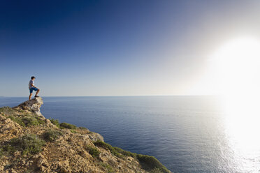 Spain, Balearic Islands, Majorca, one teenage boy standing on a rock at the cliff coast, watching the sundown - MSF004358