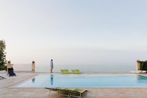 Spanien, Balearen, Mallorca, ein Jugendlicher steht auf einem sicheren Glasgeländer an einem Schwimmbad, lizenzfreies Stockfoto