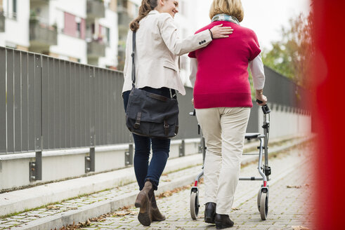 Erwachsene Enkelin, die ihrer Großmutter beim Gehen mit einer Gehhilfe auf Rädern hilft, Rückenansicht - UUF002533