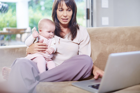 Mutter sitzt mit Baby auf der Couch und arbeitet am Laptop, lizenzfreies Stockfoto